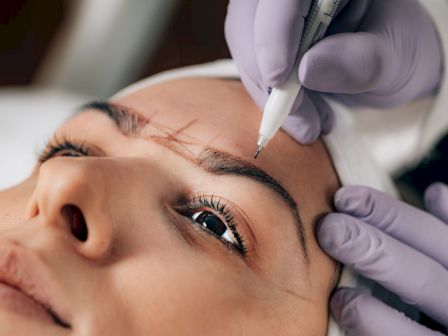 A technician in gloves uses a needle pen to perform eyebrow microblading on a client&rsquo;s face.