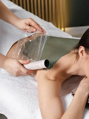 A spa therapist uses a warm wrap and massage technique while a woman lies relaxed on a treatment bed.