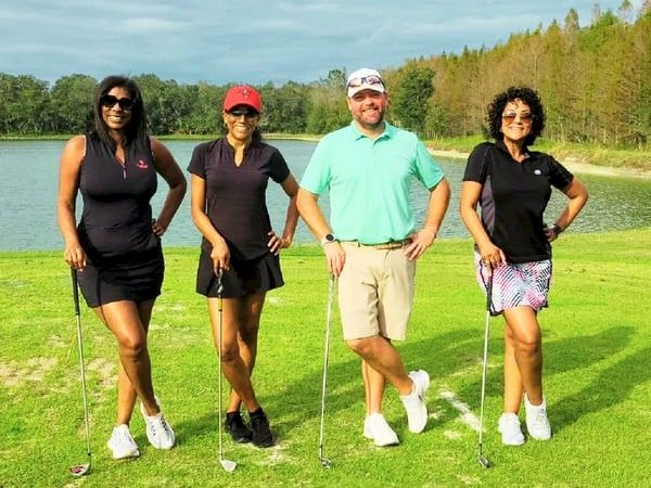A group of four people on a grassy golf course by a lake, standing with golf clubs and smiling.