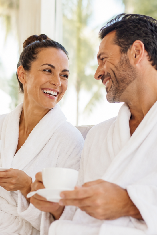 A smiling couple in white bathrobes sits on a couch, sipping tea from cups in a bright, spa-like room.