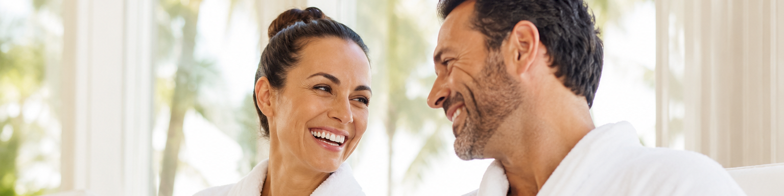A smiling couple in white bathrobes shares coffee cups on a couch, enjoying a relaxed morning together.