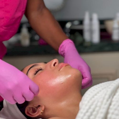 A spa worker in pink gloves performs a facial massage on a woman lying on a bed.