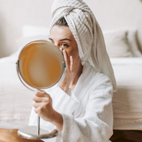 A woman in a white bathrobe and towel wrap looks into a mirror while gently applying skincare with her fingers.